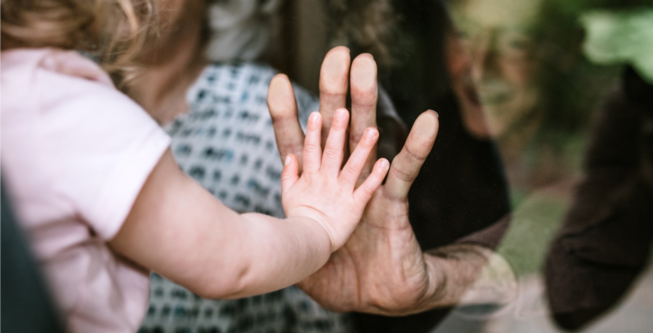 an older couple is on one side of a window, a toddler is on the other. this is a close up of their hands against the glass